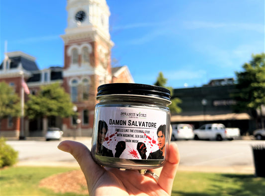 Hand holding lidded Damon Salvatore candle jar in front of a clock tower