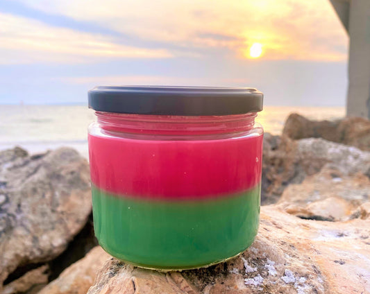 Back view of the Watermelon Sugar candle's pink-and-green layered wax jar on rocks at sunset